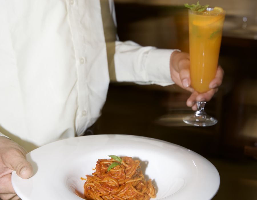 A server carrying a plate of pasta and a tall citrus spritz